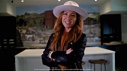 Image of a female wearing a white brimmed hat, curled red hair and a black leather jacket with her arms crossed in front of her.